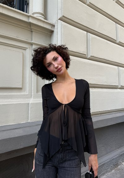 Blusa negra de manga larga con un escote en V profundo y detalle de lazo delantero, con capas de tela transparente sobre una base sólida, combinada con vaqueros oscuros.