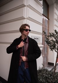 Homme portant un manteau noir par-dessus une veste en cuir bordeaux, avec des lunettes de soleil foncées, debout contre un mur blanc texturé. Plante verte à proximité.