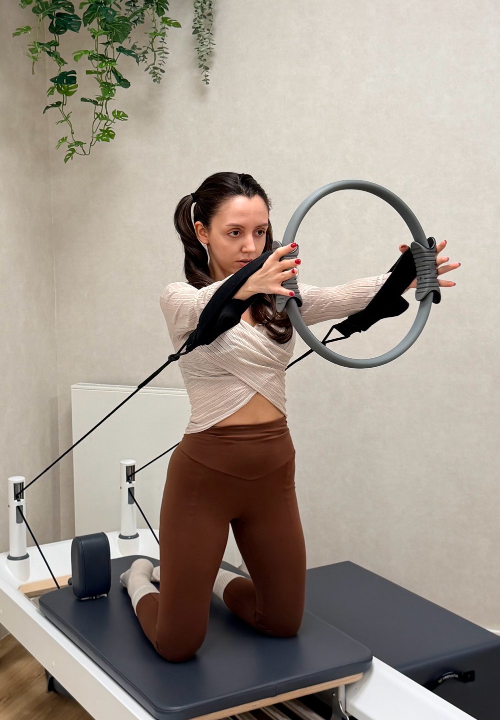 Pilates obroč s sivimi in črnimi trakovi, ki ga drži oseba, ki kleči na reformerju, oblečena v tesen kremni vrh in rjave leggice.