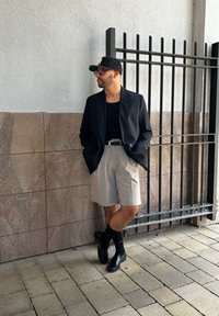 Černý blejzr přes černé tílko, světle šedé kraťasy a černé mokasíny s černými ponožkami. Doplňky zahrnují černou čepici a sluneční brýle.