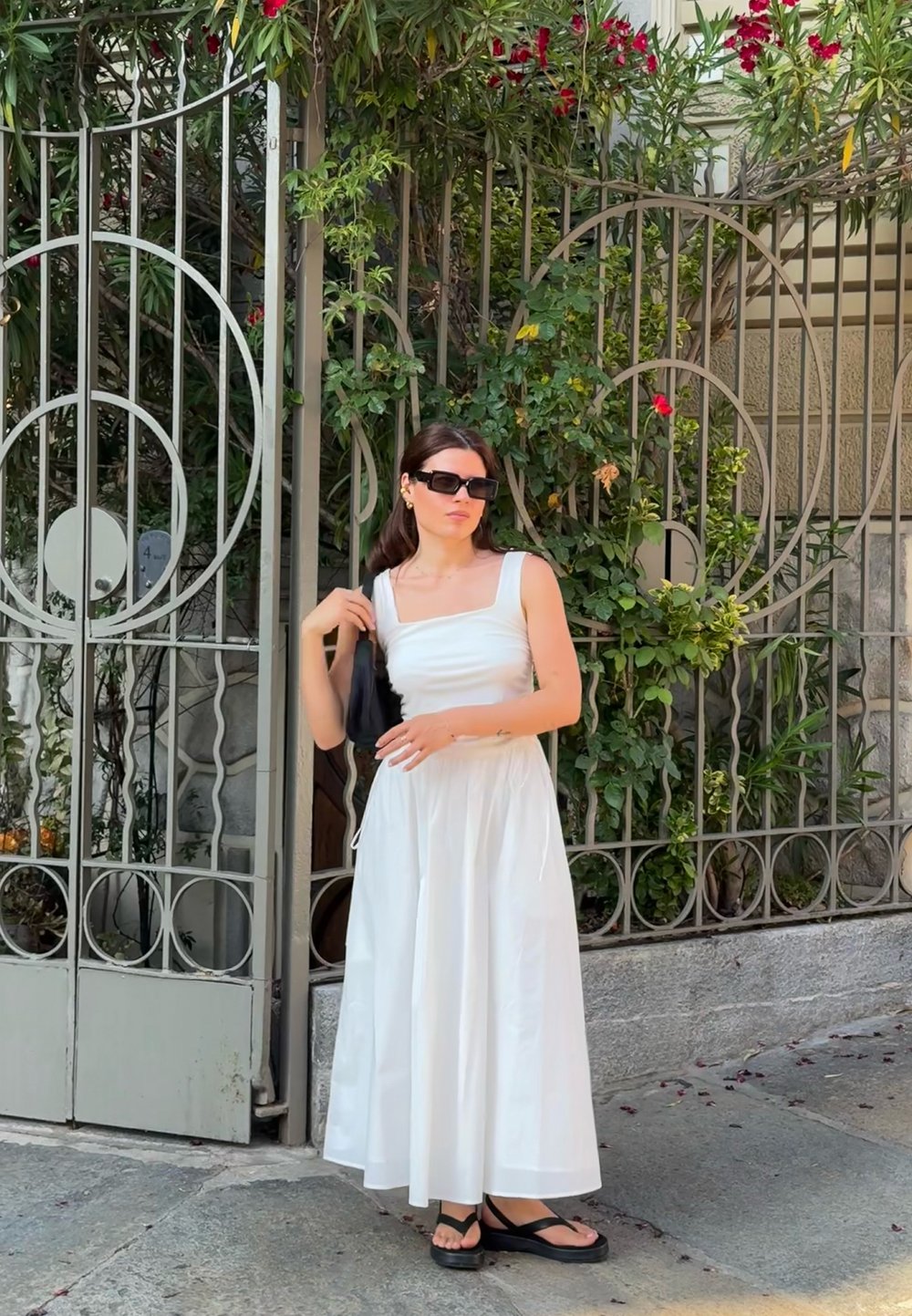 Top corto bianco senza maniche con scollo quadrato, abbinato a una lunga gonna bianca voluminosa. Una borsa nera e sandali completano l'outfit. Sfondo di fogliame verde.