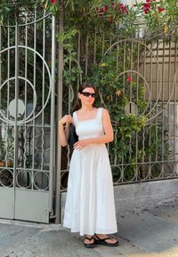 Top corto bianco senza maniche con scollo quadrato, abbinato a una lunga gonna bianca voluminosa. Una borsa nera e sandali completano l'outfit. Sfondo di fogliame verde.
