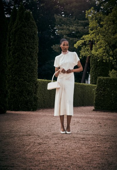 Vestido midi blanco con mangas cortas y cuello alto, que presenta patrones intrincados. Accesorios incluyen un pequeño bolso blanco y tacones.