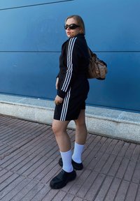 Sort atletisk outfit med hvide striber, parret med sorte sko og hvide sokker. Brun rygsæk, solbriller, kort hår, stående mod en blå væg.