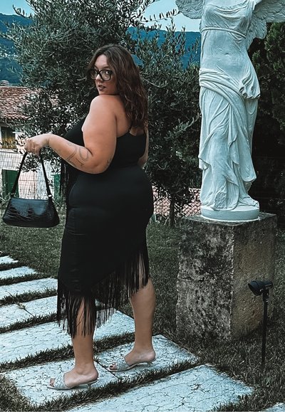 Vestido negro con flecos y una silueta ajustada, que presenta tirantes delgados. Los accesorios incluyen un pequeño bolso negro texturizado y sandalias de tacón abierto.