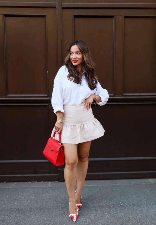 Femme aux cheveux longs portant un chemisier blanc, une jupe beige, des escarpins à fleurs, et tenant un sac à main rouge, debout contre un mur marron.