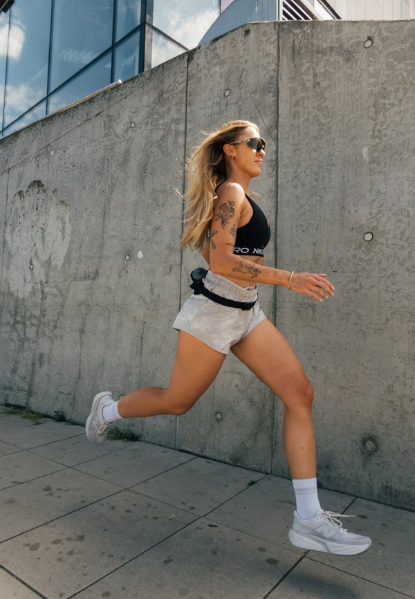Ung kvinde med tatoveringer løber udendørs ved siden af en betonvæg, iført solbriller, sportstop, shorts og sneakers på en solrig dag.