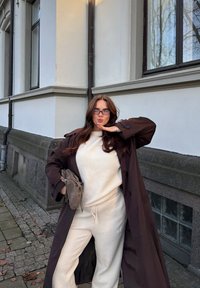 Brun trenchcoat over en kremfarget genser og matchende joggebukser, holder en brun håndveske. Briller med subtilt design, avslappet passform.