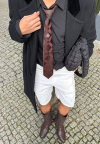 Cravatta a quadri marrone, camicia nera, pantaloncini bianchi e stivali da cowboy marroni. Soprabito nero, tiene in mano una borsa imbottita nera.