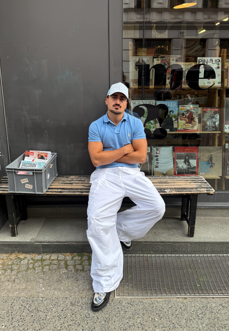Homme en polo bleu et pantalon blanc assis, les bras croisés, sur un banc devant un magasin avec des magazines en vitrine derrière lui.