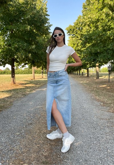 Camiseta blanca ajustada, falda maxi de denim azul claro con abertura frontal y zapatillas deportivas blancas. La escena presenta un camino flanqueado por árboles.