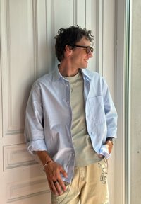 Világoskék, gombos ing hosszú ujjal egy zöld kötött felsőn; bézs nadrág hímzett részletekkel; szemüveget és kiegészítőket visel.