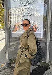 Mulher com um trench coat olival e óculos de sol pretos, carregando uma grande bolsa de palha preta, parada junto a uma paragem de autocarro com reflexo de um edifício.