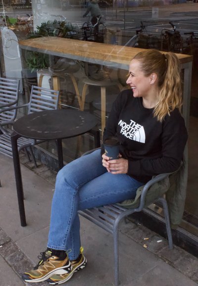 Sudadera negra con logo blanco, jeans azules y zapatillas de running marrones y amarillas. Sosteniendo un vaso de papel negro mientras está sentado en una mesa negra.
