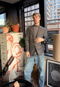 Νεαρό άτομο με γκρι σακάκι και τζιν στέκεται μέσα σε εσωτερικό χώρο δίπλα σε ένα αρχείο με πορτοκαλί γκράφιτι φλαμίνγκο και γλάστρες κοντά σε ένα παράθυρο.