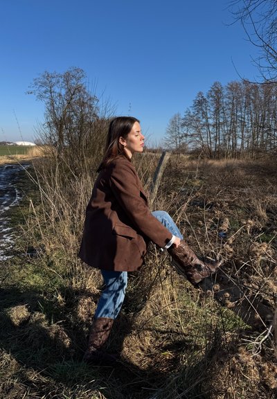 Blazer de ante marrón, jeans azules y botas altas marrones usadas por una persona de pie al aire libre. La escena incluye hierba seca y un cielo azul claro.