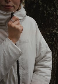 Hvid, isoleret jakke med høj krave og lynlås, med et diskret logo. Glat tekstur og afslappet pasform, båret med en strikket hat.