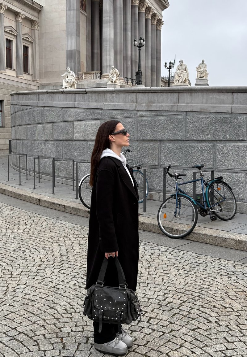Manteau noir, sweat à capuche gris, baskets argentées. Tient un sac noir avec des clous. Fond en pavés gris clair avec des bicyclettes et un bâtiment en pierre.