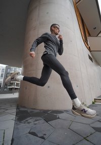 Homme athlétique en haut à manches longues sombre et leggings noirs en train de courir. Il porte des baskets grises et blanches avec un accent violet. Fond de mur en béton.