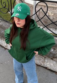 Sweat à capuche vert avec une poche sur le devant et des manches longues, associé à un jean évasé bleu clair et une casquette de baseball verte assortie.