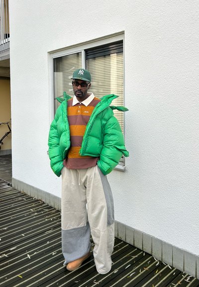 Chaqueta acolchada verde brillante, camiseta tipo polo a rayas marrón y naranja, pantalones de pierna ancha gris y beige, y zapatos color tostado, contra un fondo de pared blanca.