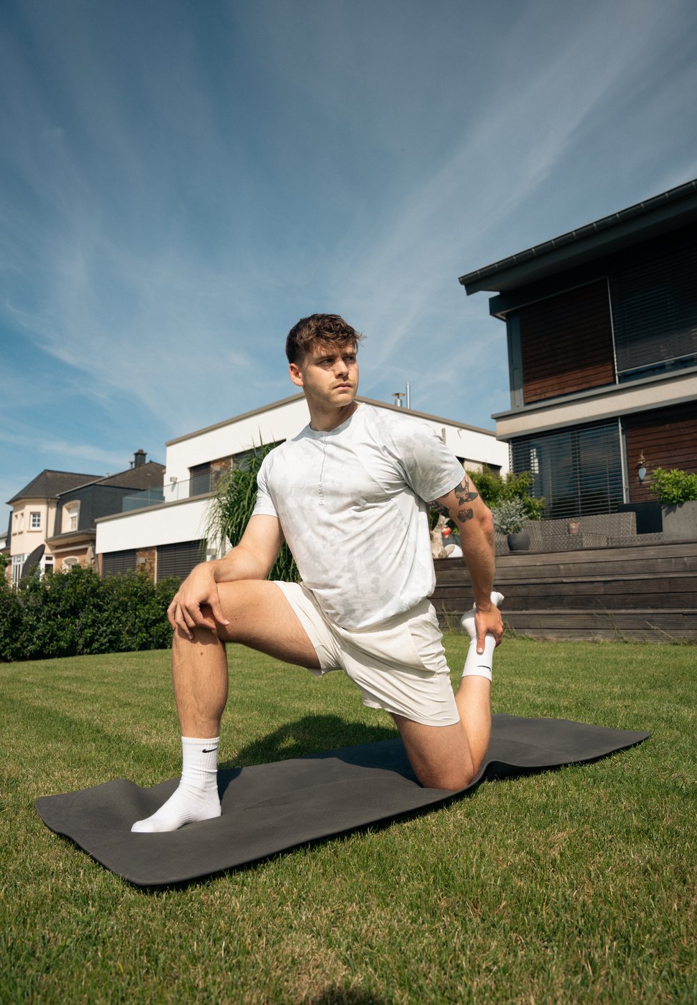 Homem a alongar-se em um tapete de yoga cinza escuro em uma área gramada, vestindo uma camisa cinza claro camuflada e calções brancos, com meias brancas e uma tatuagem.