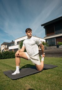 Homem a alongar-se em um tapete de yoga cinza escuro em uma área gramada, vestindo uma camisa cinza claro camuflada e calções brancos, com meias brancas e uma tatuagem.