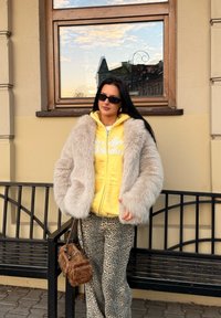 Casaco de pele sintética com textura cinza, hoodie amarelo com fecho, calças bege com padrão, óculos de sol e uma pequena bolsa castanha. Sentada junto a um banco preto.