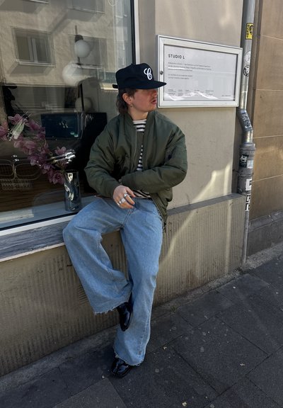 Chaqueta bomber verde, camisa de rayas, pantalones anchos de mezclilla de color azul claro, zapatos negros y gorra con un logo. Los accesorios incluyen múltiples anillos.