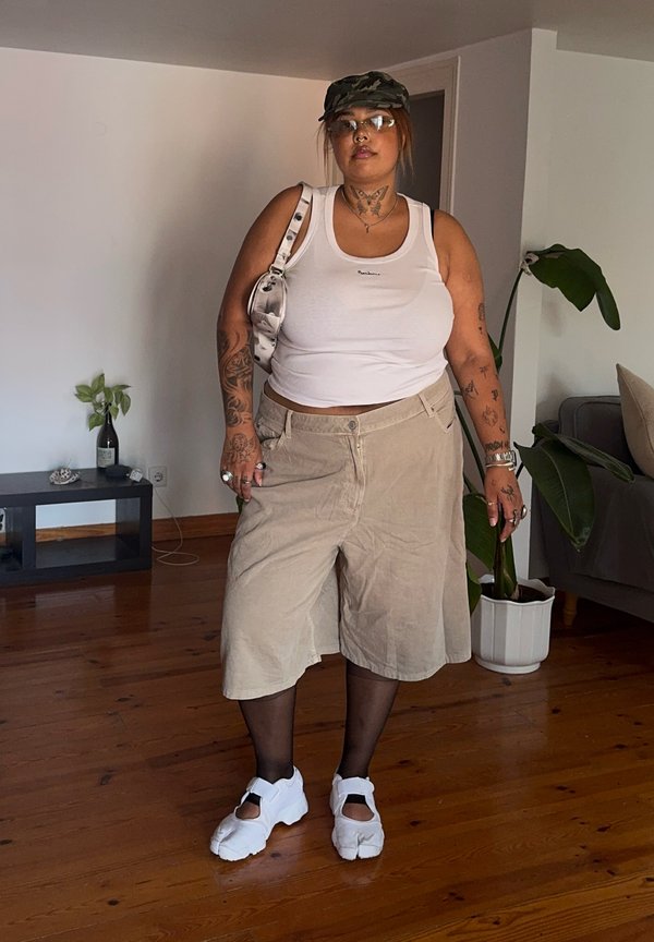 Persoon draagt een wit tanktop, beige lange shorts, zwarte panty's, witte sandalen, camouflagpet, bril en draagt een witte schoudertas binnen.