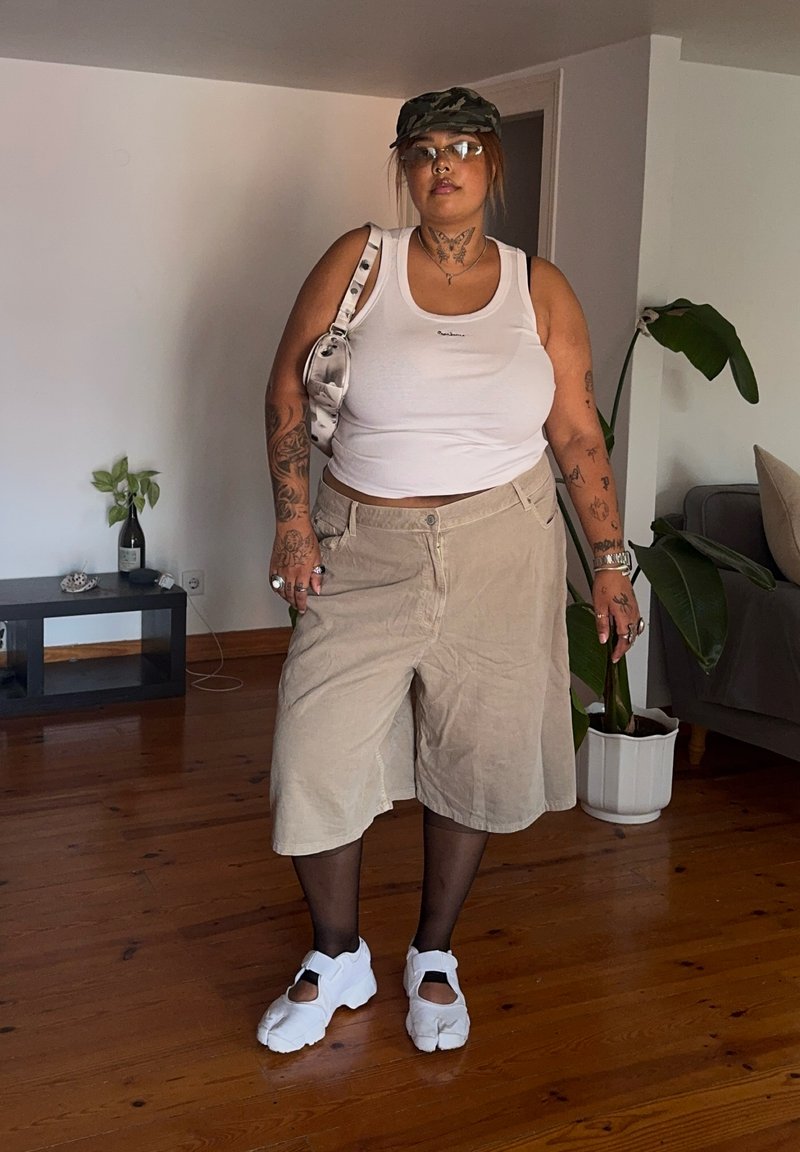 Personne portant un débardeur blanc, un short long beige, des collants noirs, des sandales blanches, une casquette camouflage, des lunettes et portant un sac à bandoulière blanc à l'intérieur.