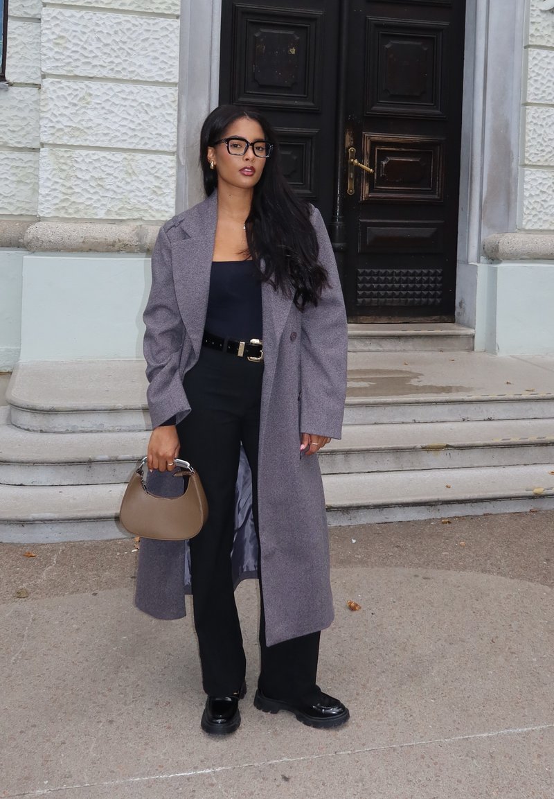 Γκρίζο υπερμεγέθες παλτό, μαύρη μπλούζα και μαύρο παντελόνι. Κρατάει μια καφέ τσάντα χειρός. Γυαλιστερά μαύρα παπούτσια, μακριά σκούρα μαλλιά και γυαλιά με μαύρο σκελετό.