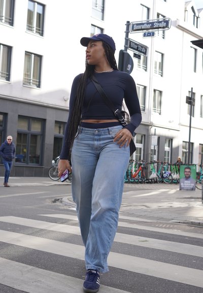 Una persona lleva un top de manga larga azul marino y jeans holgados azul claro, sostiene un teléfono inteligente, camina por un paso de peatones con una gorra y zapatillas.