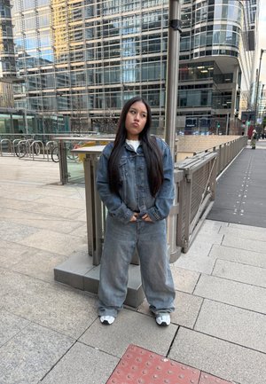 Chaqueta de denim y jeans holgados en azul desgastado. Camiseta blanca debajo y zapatillas grises. De pie sobre un pavimento de piedra con edificios modernos detrás.