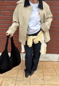 Personne portant une veste beige, un t-shirt blanc, un jean foncé, des chaussures noires à bouts carrés, tenant un grand sac noir, debout contre un mur en briques rouges.