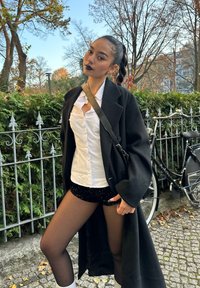 Zwart oversized mantel over een wit overhemd met knopen en zwarte kortebroek met pailletten. Ze draagt zwarte sheer maillots en witte sokken, met haar in een vlecht.