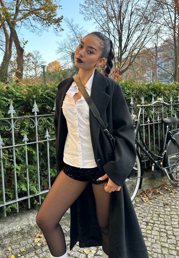 Svart oversized kappa över en vit skjorta med knappar och svarta paljettshorts. Hon bär genomskinliga svarta strumpbyxor och vita strumpor, med håret flätat.