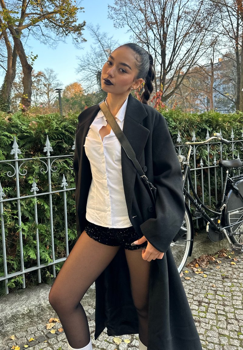 Schwarzer übergroßer Mantel über einem weißen Hemd mit Knöpfen und schwarzen Pailletten-Shorts. Sie trägt transparente schwarze Strumpfhosen und weiße Socken, das Haar ist zu einem Zopf geflochten.