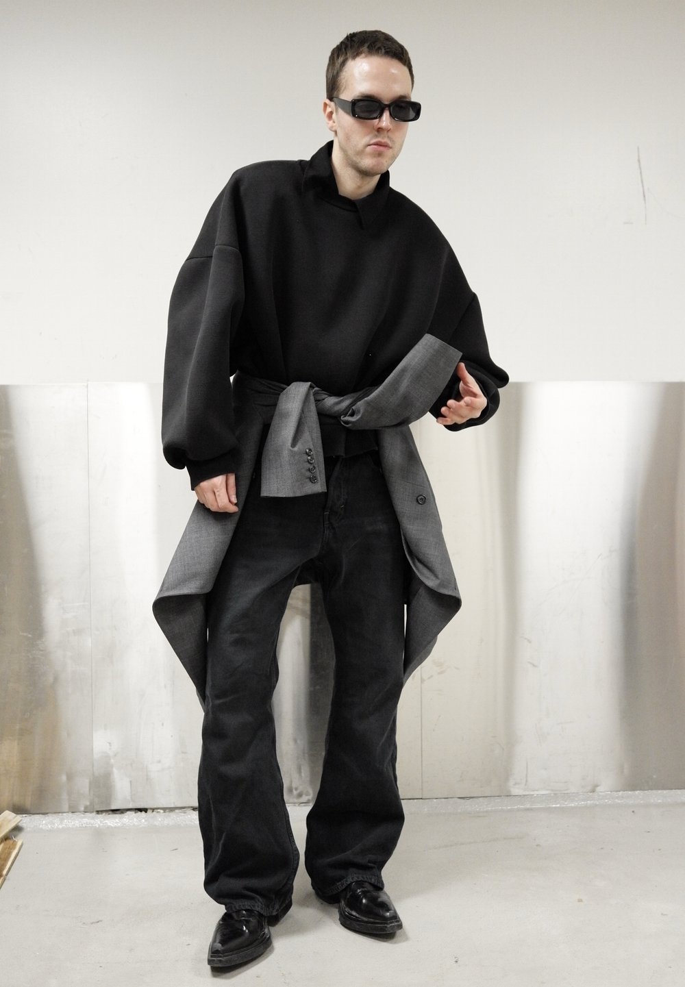 Sweat-shirt noir surdimensionné avec un col montant, tissu gris porté en guise de ceinture, associé à un jean noir ample et des chaussures noires.