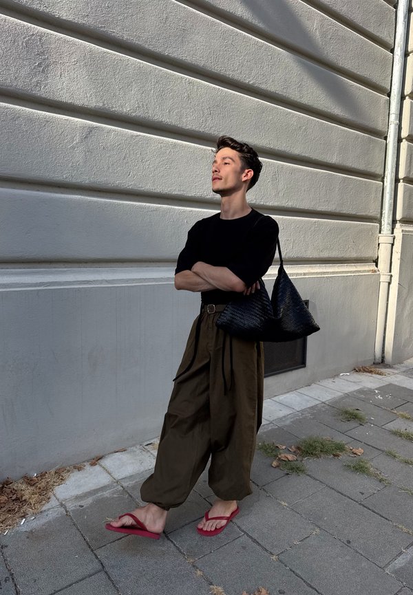 Un homme portant une chemise noire, un pantalon marron ample et des sandales rouges se tient les bras croisés près d’un mur en pierre grise, avec un sac noir tressé.