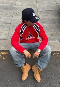 Osoba oblečená v červenej mikine Adidas, modrých džínsoch, hnedej obuvi a čiernej šiltovke sedí na obrubníku a pozerá sa do strany na povrchu chodníka.