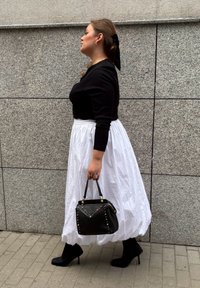 Mujer caminando al lado de una pared de piedra gris, vistiendo una blusa negra, una falda midi blanca, botas de tacón negras y sosteniendo un bolso negro con tachuelas de colores.