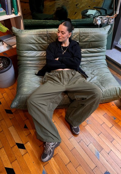 Pantalones verdes de gran tamaño con cordón, sudadera negra y zapatillas de deporte con estampado sobre un sofá de cuero verde texturizado en un interior moderno.