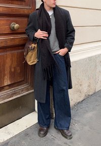 Personne portant un manteau gris foncé, une écharpe noire, un pull gris, un jean large bleu foncé, des chaussures marron, et portant un sac à bandoulière beige et marron.