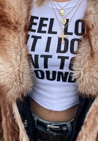 Cămașă albă scurtă cu text negru, purtată sub o haină din blană sintetică bej; mai multe coliere din aur cu pandantive. Blugi albaștri cu o curea decorativă.