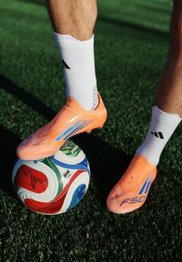 Άτομο που φοράει πορτοκαλί ποδοσφαιρικά παπούτσια και λευκές κάλτσες ελέγχει μια πολύχρωμη ποδοσφαιρική μπάλα σε πράσινο τεχνητό χλοοτάπητα.