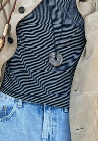 Μπλούζα με μαύρες και άσπρες ρίγες κάτω από ένα μπεζ σουέτ μπουφάν. Ένα στρογγυλό κρεμαστό από πέτρα κρέμεται από μια μαύρη κορδόνι. Ορατά ελαφριά μπλε τζιν παντελόνια.