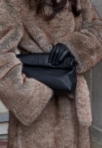 Manteau en fausse fourrure dans des tons bruns avec une texture douce. Pochette en cuir noir tenue avec des gants en cuir noir, mettant en avant des lignes épurées et un contraste marqué.