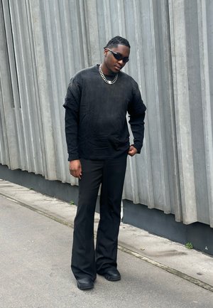 Hombre vestido con ropa completamente negra, gafas de sol y cadena plateada, de pie contra una pared gris texturizada en una acera urbana.