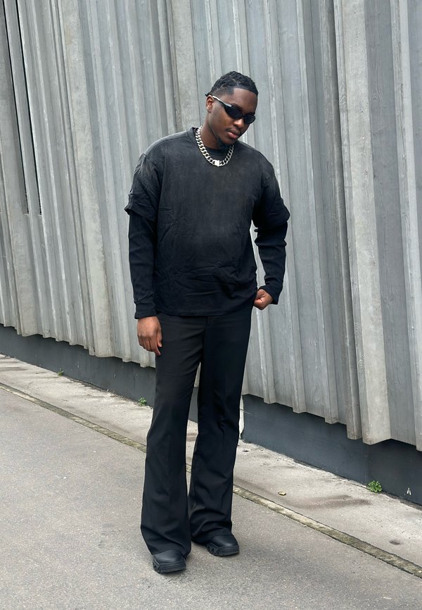 Bărbat îmbrăcat în negru complet, purtând ochelari de soare și un lanț argintiu, stă în fața unui perete gri texturat pe trotuarul urban.